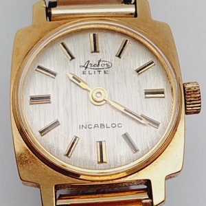 May include: A gold-toned wristwatch with a square face. The watch face is white with black markings and the text "Arcturus Elite" and "Incabloc".