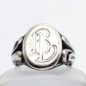 Può includere: Anello con sigillo in argento con una faccia ovale con la lettera "B" in un carattere serif. L'anello ha un design ornato sulle spalle e una finitura lucida. Un gioiello classico.