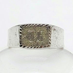 May include: A silver-toned ring with a rectangular face. The face has a textured, gold-colored center with an ornate initial design. The ring has a rope-like border around the face and a smooth, wide band.