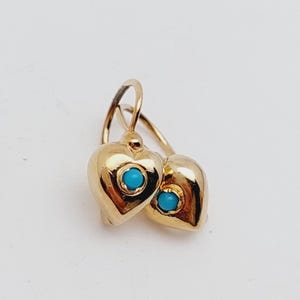 May include: A pair of gold-colored heart-shaped earrings. Each earring features a small, round, turquoise-colored stone set in the center of the heart. The earrings have a simple, elegant design with a hook closure.