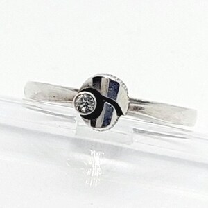 Puede incluir: Un anillo de tono plateado con una banda lisa y pulida y un adorno circular central. El adorno presenta una pequeña piedra redonda facetada y transparente a un lado, contrastada por una forma oscura, curva y abstracta en la superficie.