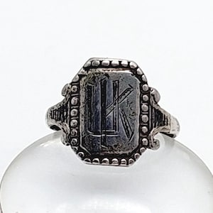 May include: A silver-toned ring with a rectangular face featuring the engraved initials "LK". The ring has an ornate design with beaded and decorative elements around the face and shoulders. The ring is set on a clear, rounded surface.
