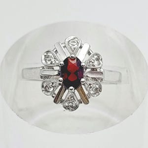 Puede incluir: Anillo de plata con una piedra preciosa central ovalada de color rojo intenso. La piedra preciosa está rodeada por un engaste decorativo con piedras preciosas más pequeñas y claras. El anillo tiene un diseño clásico y elegante.