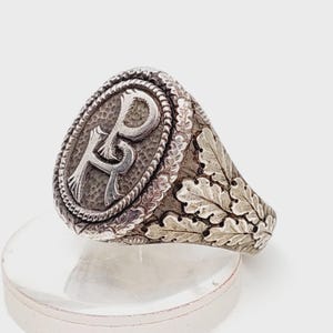 May include: Silver signet ring with an oval face featuring the intertwined letters "R" and "P" within a rope-like border. The ring's band is adorned with detailed oak leaf designs, creating a vintage aesthetic.