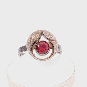 May include: A silver ring with a textured, circular design surrounding a round, red gemstone. The ring has a simple band and a unique, artistic style. The gemstone is a vibrant red color.