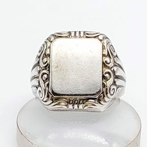 Può includere: Un anello con sigillo d'argento con una faccia rettangolare. L'anello presenta volute ornate e un centro lucido e vuoto. L'anello è esposto su una superficie bianca, mettendo in risalto il suo design intricato e la lucentezza metallica.