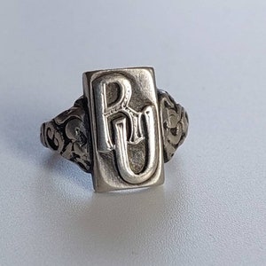 Könnte beinhalten: Ein silberner Ring mit einem rechteckigen Band, auf dem die Initialen "RU" in einer stilisierten Schriftart graviert sind. Das Band ist mit einem floralen Muster verziert.