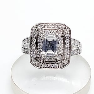 Könnte beinhalten: Ein silberner Ring mit einem großen, rechteckigen, klaren Edelstein in der Mitte, umgeben von einem doppelten Halo aus kleineren, runden, klaren Steinen. Zusätzliche klare Steine ​​sind auf dem Band angebracht. Der Ring wird auf einer weißen Oberfläche präsentiert.