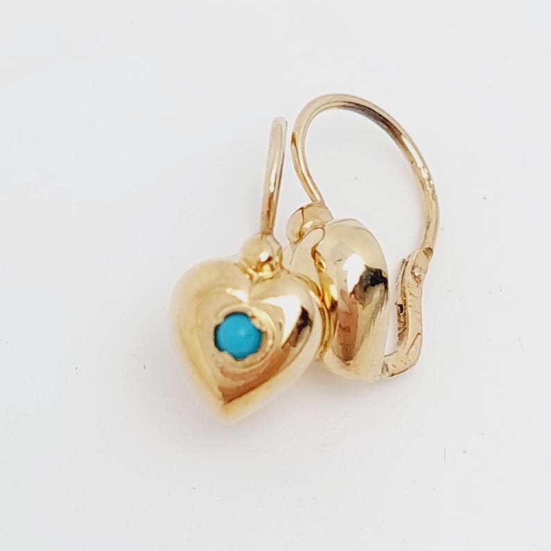 May include: A pair of gold heart-shaped earrings with a small turquoise stone in the center of each heart. The earrings have a lever back closure and are displayed against a white background.