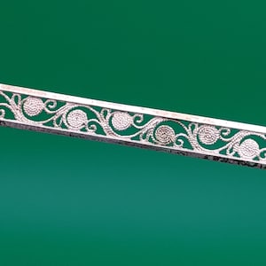 May include: A silver filigree bar brooch with a delicate floral design. The brooch is made of intricate, interwoven silver wire that creates a pattern of leaves and swirls.