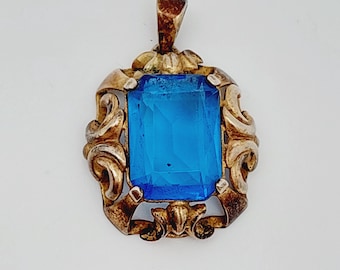 May include: Vintage pendant featuring a large, rectangular, bright blue gemstone set in a decorative, ornate gold-toned metal frame. The pendant has a bail for attaching to a chain. The design is reminiscent of antique jewelry.