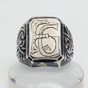 Puede incluir: Un anillo de sello plateado con una cara rectangular. La cara presenta una letra "G" grabada en una fuente decorativa. El anillo tiene adornos de volutas en los lados.