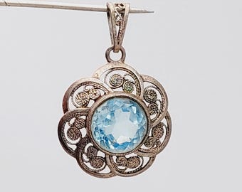 May include: A silver pendant with a filigree flower design. The pendant features a round, light blue gemstone at its center. The filigree work creates a delicate, openwork pattern around the gemstone, and the pendant is suspended from a small loop.