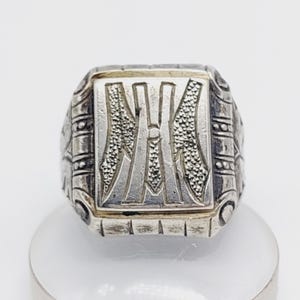 May include: A silver-toned ring with a rectangular face featuring an embossed design. The ring's face has a central letter "H" flanked by textured patterns. The band has a detailed, geometric design. The ring is displayed on a white surface.