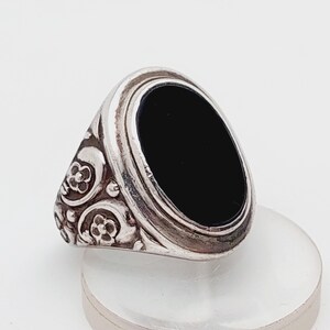 Puede incluir: Anillo de plata con una piedra negra ovalada. El anillo tiene un diseño ornamental en los laterales con motivos florales. El anillo está sobre una superficie clara y redondeada.