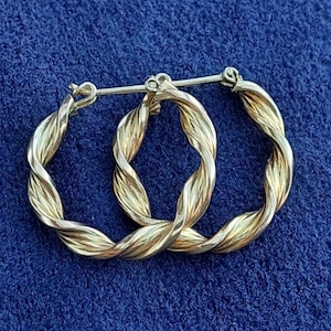 Puede incluir: Un par de pendientes de aro dorados con un dise&ntilde;o trenzado. Los pendientes est&aacute;n hechos de metal y tienen un acabado brillante.