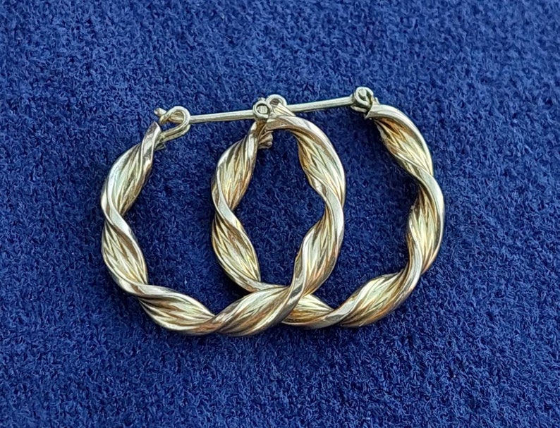 Puede incluir: Un par de pendientes de aro dorados con un dise&ntilde;o trenzado. Los pendientes est&aacute;n hechos de metal y tienen un acabado brillante.