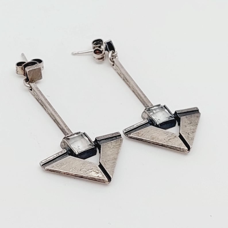 Puede incluir: Un par de pendientes de plata con un dise&ntilde;o geom&eacute;trico. Cada pendiente presenta un cristal transparente en el centro de una forma triangular.