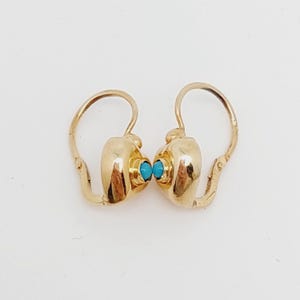 May include: A pair of gold-colored earrings with a turquoise stone in the center of each earring. The earrings have a hook-style closure. The earrings are displayed on a white background.
