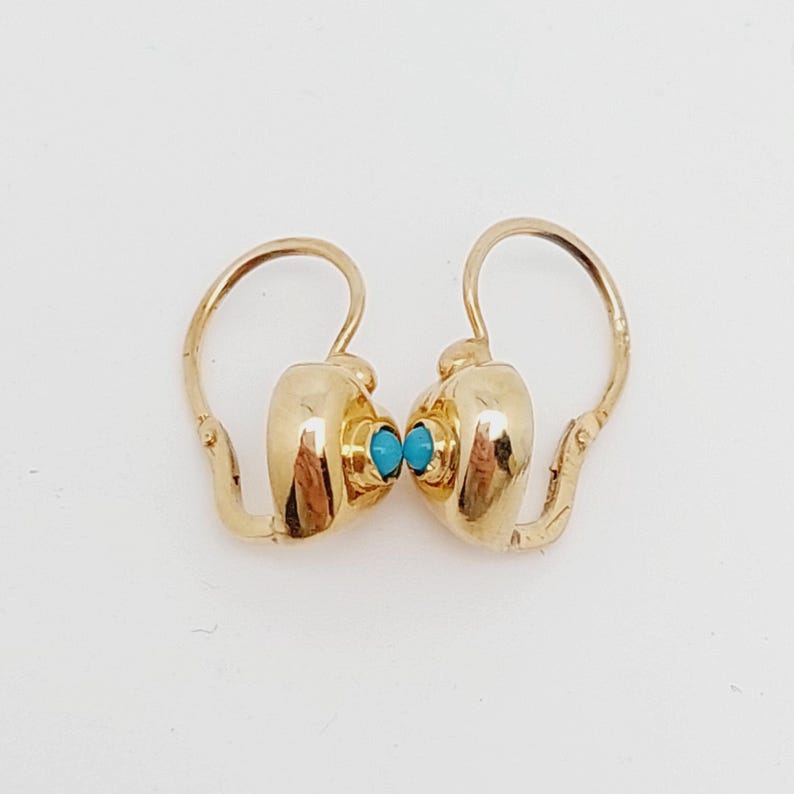 May include: A pair of gold-colored earrings with a turquoise stone in the center of each earring. The earrings have a hook-style closure. The earrings are displayed on a white background.