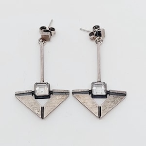 Puede incluir: Un par de pendientes de plata con un dise&ntilde;o geom&eacute;trico. Cada pendiente presenta una piedra de cristal transparente de forma cuadrada, engastada en un marco de plata. Los pendientes tienen una varilla larga y fina de plata que cuelga de un peque&ntilde;o gancho de plata.