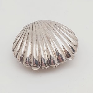 Könnte beinhalten: Ein silberfarbenes, muschelförmiges Dekorationsobjekt. Das Objekt hat eine geriffelte Textur und einen gewellten Rand. Das Objekt ist wahrscheinlich aus Metall und vor einem weißen Hintergrund platziert. Das Objekt ist ein dekorativer Artikel.
