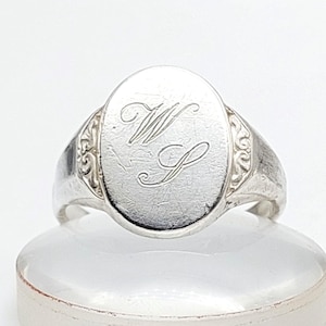 May include: Silver signet ring with an oval face engraved with the letters "W" and "S" in a script font. The ring has decorative detailing on the shoulders and a polished finish.