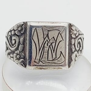 May include: A silver signet ring with a rectangular face. The face features an engraved monogram, with floral and scrollwork details on the sides. The ring is set against a white background.