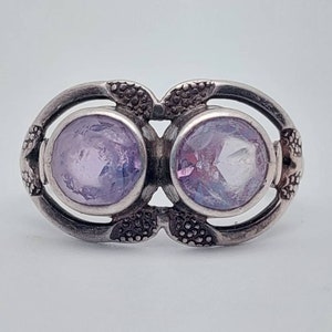 May include: A silver ring with two round, faceted, light purple gemstones set in a decorative, ornate design.