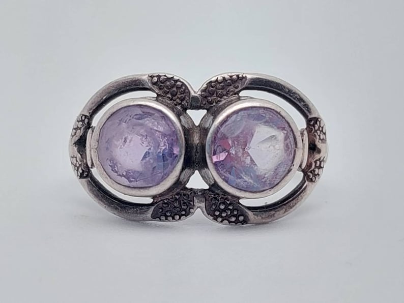 May include: A silver ring with two round, faceted, light purple gemstones set in a decorative, ornate design.