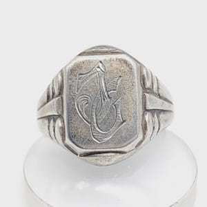 May include: A silver-toned ring with a rectangular face featuring an engraved monogram. The ring has a detailed band with vertical lines on each side of the face. The ring is displayed on a white surface.