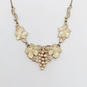 May include: A gold tone necklace with a grape cluster pendant. The pendant features a cluster of faux pearls and intricate leaf designs.