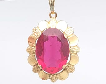 May include: A gold pendant with an oval, magenta-colored gemstone. The gemstone is set in a gold frame with a floral design. The pendant has a small loop at the top for a chain.