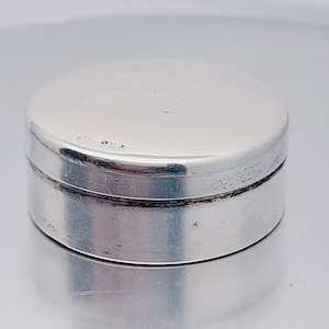 Può includere: Una scatola rotonda in argento per gioielli con una superficie liscia e lucida. Il coperchio ha una piccola iscrizione che dice "925 STERLING".