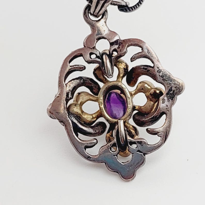 Puede incluir: Un colgante de plata y oro con una amatista p&uacute;rpura en el centro. El colgante tiene un dise&ntilde;o de filigrana y est&aacute; unido a una cadena de plata.