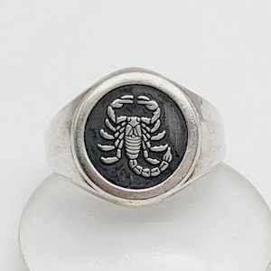 Könnte beinhalten: Ein silberner Siegelring mit einem detaillierten Skorpion-Emblem auf dunklem, kreisförmigem Hintergrund. Der Ring hat ein poliertes, abgerundetes Band und zeigt ein Tierkreis- oder astrologisches Thema.