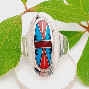 Op de afbeelding: Ovale zilveren ring met een kleurrijk inlegontwerp. De inleg heeft een centrale rode band, geflankeerd door blauwe en rode driehoeken en zwarte accenten. De ring staat tegen een witte achtergrond met groene bladeren.