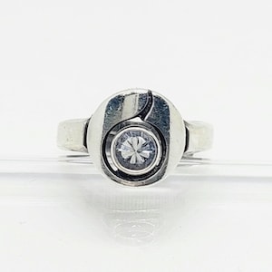 Puede incluir: Un anillo de tono plateado que presenta una pieza central circular con acabado pulido y una única piedra transparente de corte redondo engastada en el centro. La banda de metal tiene una construcción sólida y lisa con un diseño curvo.