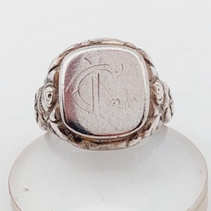 May include: A silver signet ring with a rectangular face featuring an engraved monogram. The ring has a detailed floral design around the band. The ring is displayed on a clear, rounded surface.