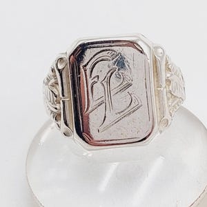 May include: Silver signet ring with a rectangular face featuring an engraved monogram. The ring has decorative floral and leaf detailing on the shoulders. The ring is displayed on a clear, rounded surface.