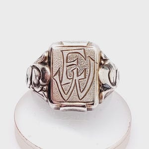 May include: A silver signet ring with an engraved initial "EW" on the face. The ring has a decorative design on the sides.