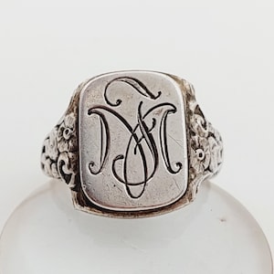 Puede incluir: Un anillo de sello de plata con un monograma grabado. La cara rectangular del anillo presenta un monograma de escritura adornado, probablemente iniciales. La banda está detallada con diseños florales y de volutas, agregando una estética vintage.