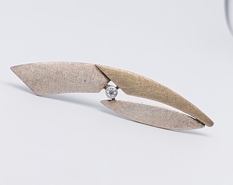 May include: A silver and gold abstract brooch with a single clear gemstone in the center.
