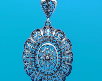 May include: A silver filigree pendant with an oval, floral design. The pendant is suspended from a delicate chain against a blue background. The intricate openwork design features swirling patterns and a heart-shaped bail.