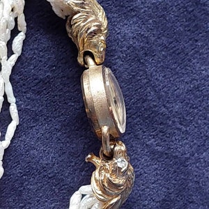 May include: A gold-tone wristwatch with a white pearl bracelet. The watch face is round and has a gold-tone bezel. The watch is adorned with two lion head clasps.