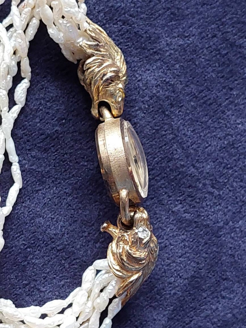 May include: A gold-tone wristwatch with a white pearl bracelet. The watch face is round and has a gold-tone bezel. The watch is adorned with two lion head clasps.