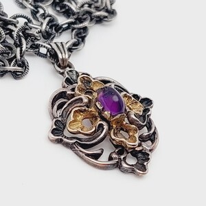 Puede incluir: Un collar de cadena de plata con un colgante que presenta una piedra preciosa p&uacute;rpura rodeada de filigrana dorada y plateada.