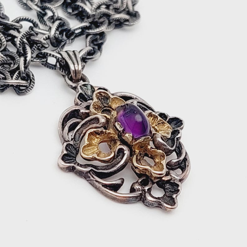 Puede incluir: Un collar de cadena de plata con un colgante que presenta una piedra preciosa p&uacute;rpura rodeada de filigrana dorada y plateada.