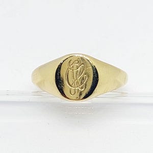 May include: A gold signet ring featuring a smooth, polished surface with an oval face. The center of the oval face is engraved with an intricate, stylized monogram consisting of intertwined cursive letters that appear to be the characters C and L.
