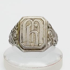 May include: Silver-toned signet ring featuring the letter "H" in a rectangular, engraved design. The ring has ornate scrollwork on the sides and is displayed on a clear stand. A classic piece of jewelry.